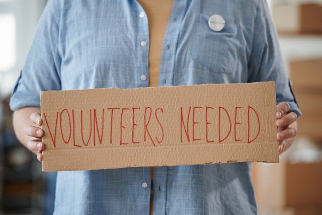Person holding a cardboard sign that reads volunteers needed