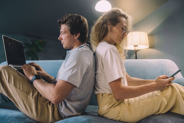 Two people sitting back to back, one using a laptop and the other using a phone, representing online chat and digital communication